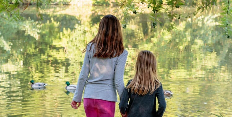 Older and younger sister holding hands and watching lake in autumn