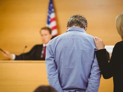 Man wearing blue shirt with hands behind his back and head down and judge about to bang gavel on sounding block in background