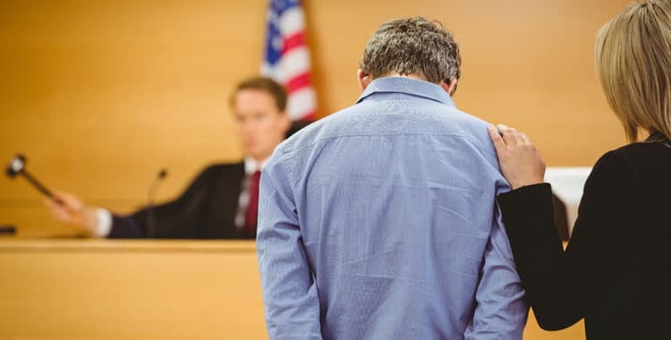 Man wearing blue shirt with hands behind his back and head down and judge about to bang gavel on sounding block in background