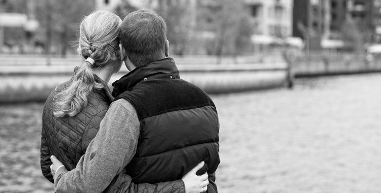grayscale photo of the backs of a young couple wrapping their arms around each other and looking at a river