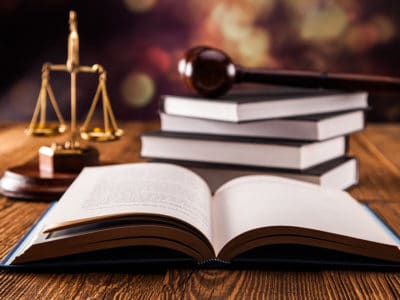Open book in foreground with gold scales of justice and gavel resting on pile of books in background