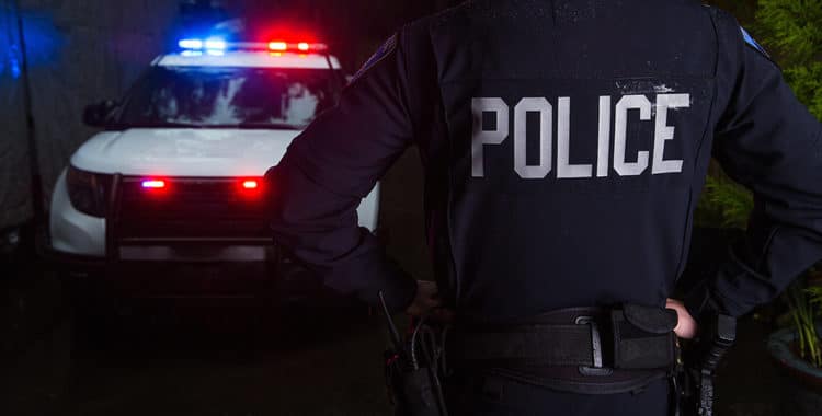 police officer standing in foreground with white police SUV with lights flashing in background