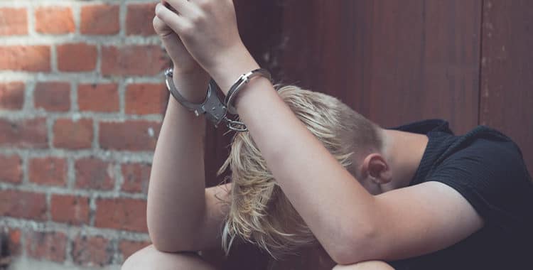 Caucasian teen sitting on ground with head down and elbows on knees wearing handcuffs