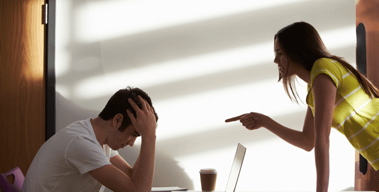 Young college man sitting at desk with hands on head while young college woman points her finger at him